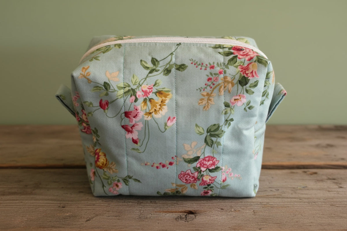 A square fabric cosmetic pouch with a floral print on a light blue-green background, featuring pink and yellow flowers with green leaves, and a white zipper at the top. It is placed rustic board.
