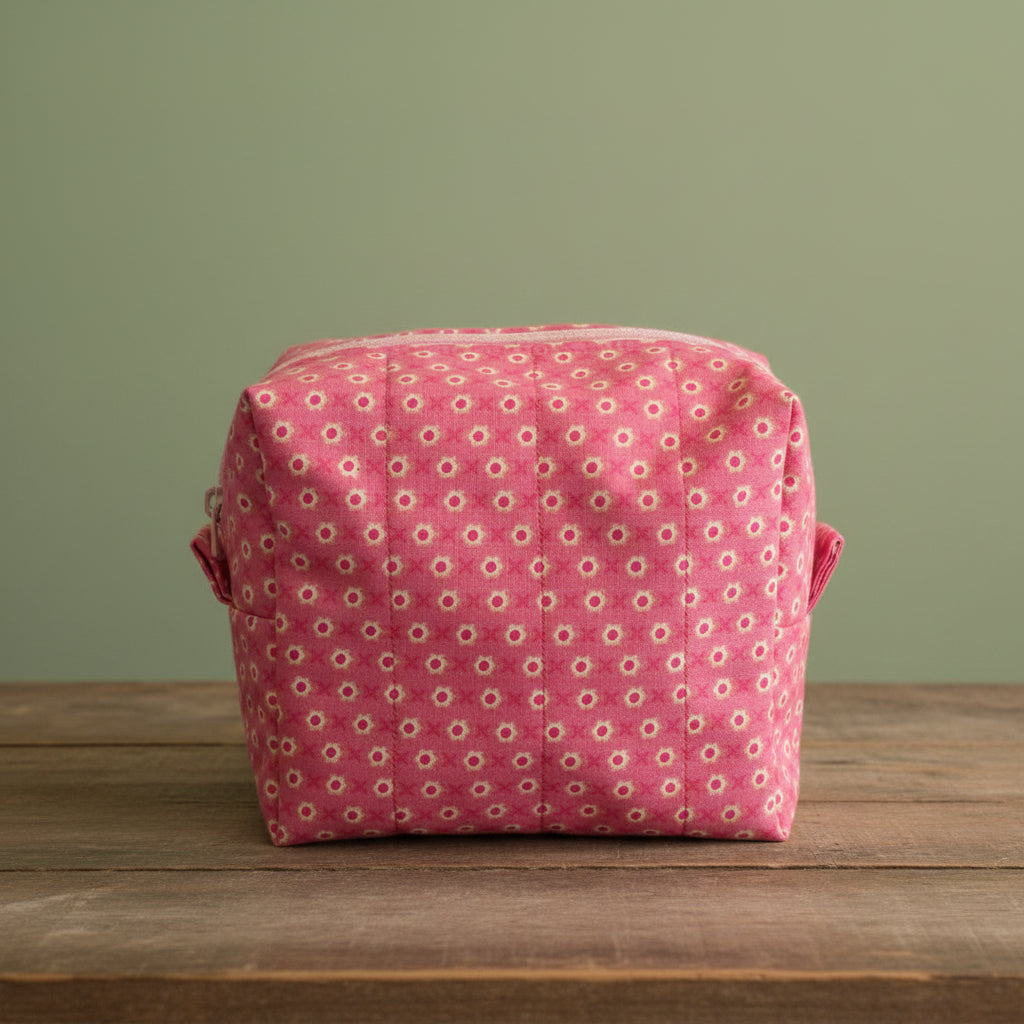 A pink fabric zippered pouch with a white and hot pink polka dot pattern, resting on a rustic board surface against a sage green wall.
