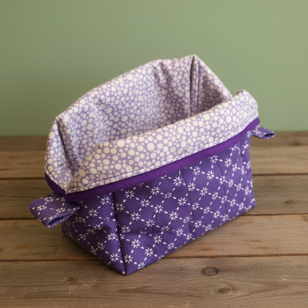 A fabric mini cosmetic bag with a purple exterior with a white flower and lattice patterned exterior and a light purple with a white polka dotted patterned interior, resting on a rustic board.