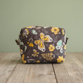 A square cosmetic bag with sage green background and a colorful butterfly and floral pattern in yellow, white, and light green, resting on a rustic board