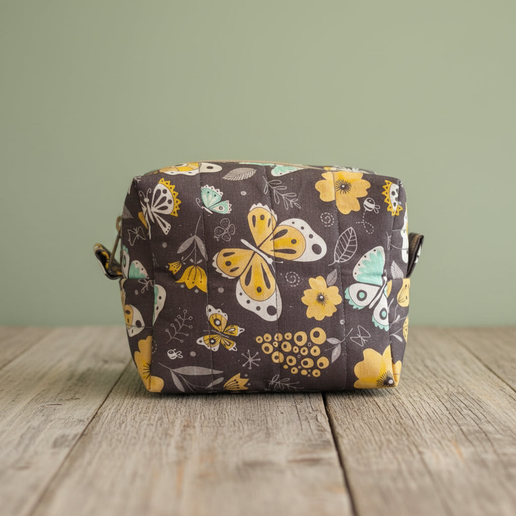 A square cosmetic bag with sage green background and a colorful butterfly and floral pattern in yellow, white, and light green, resting on a rustic board