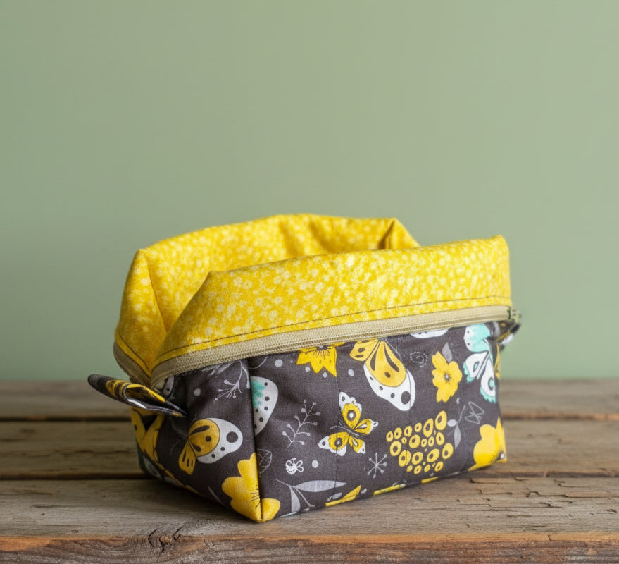 A fabric mini cosmetic bag with a yellow patterned interior and a dark gray exterior featuring a yellow and white, yellow, and light blue butterfly pattern sitting on a rustic board.