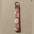 A fabric wristlet keychain with a dusty rose-pink background and a white daisy floral pattern, featuring a silver metal clasp and split ring