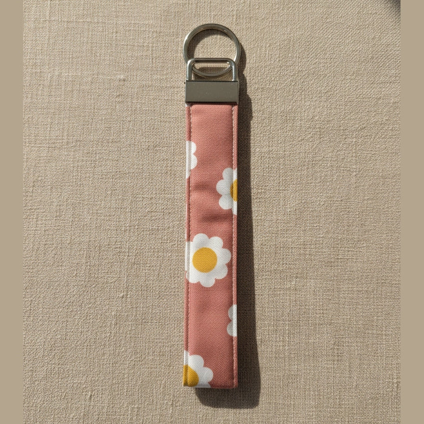 A fabric wristlet keychain with a dusty rose-pink background and a white daisy floral pattern, featuring a silver metal clasp and split ring