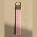 A pink fabric keychain strap with small gold star details and a silver metal split ring, shown against warm linen fabric.