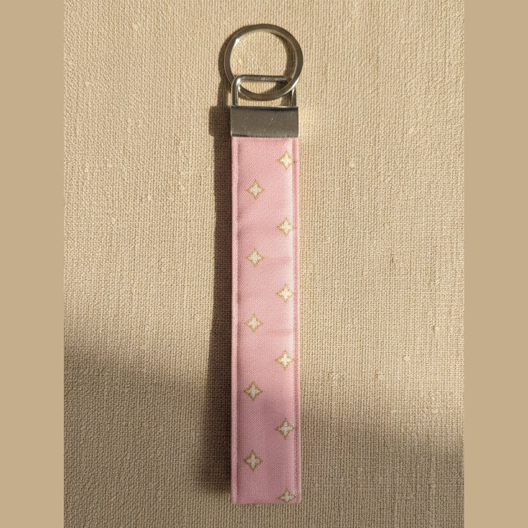 A pink fabric keychain strap with small gold star details and a silver metal split ring, shown against warm linen fabric.
