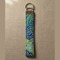 A fabric keychain pouch with a blue, green, and yellow paisley pattern, attached to a silver metal split ring, shown against a warm linen background