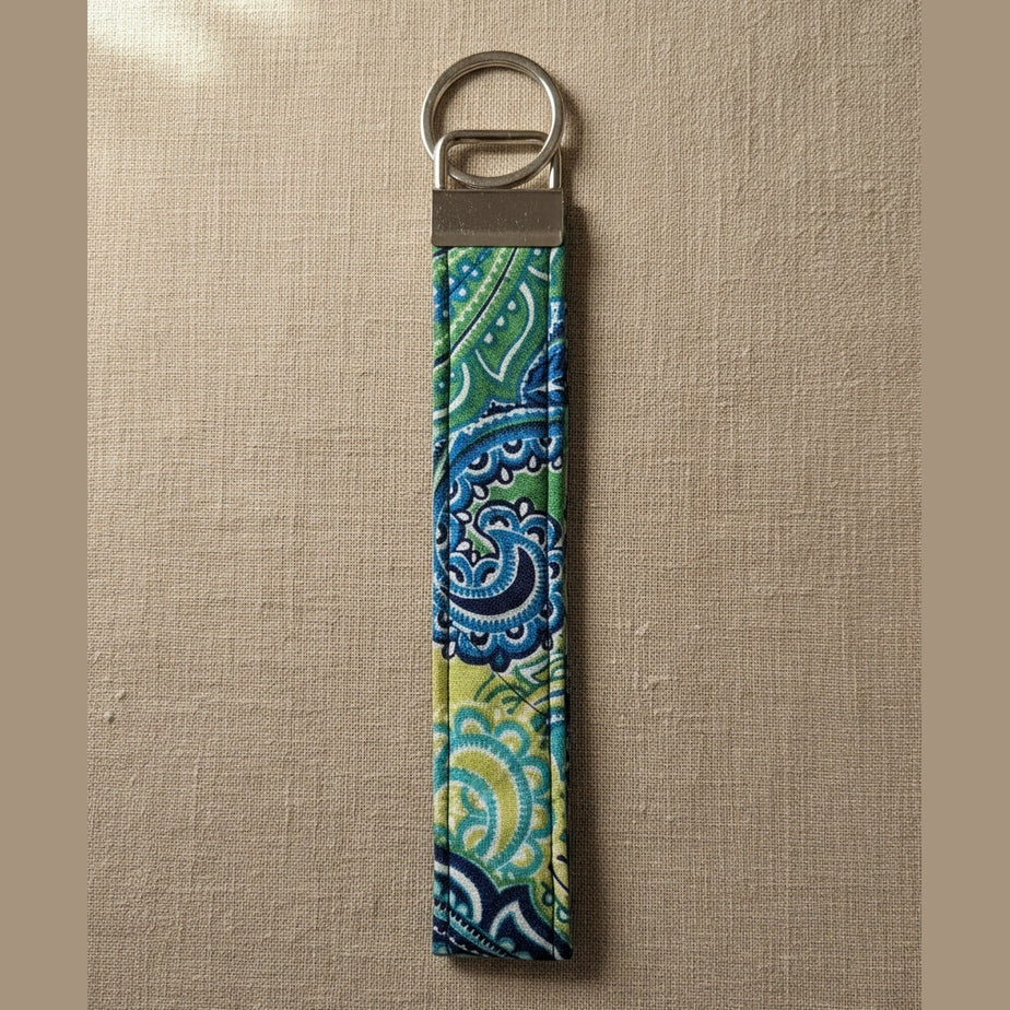 A fabric keychain pouch with a blue, green, and yellow paisley pattern, attached to a silver metal split ring, shown against a warm linen background