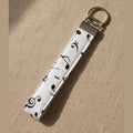 A fabric wristlet keychain with a black musical note pattern on a white background, featuring a silver metal clasp and keyring, shown against a warm linen background.