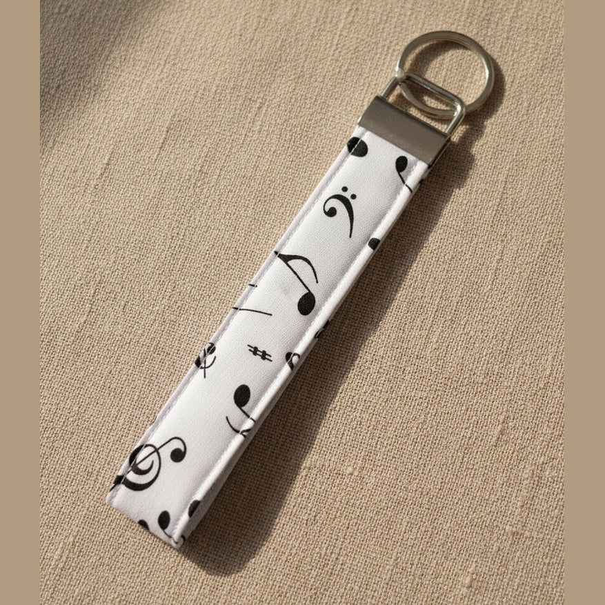 A fabric wristlet keychain with a black musical note pattern on a white background, featuring a silver metal clasp and keyring, shown against a warm linen background.