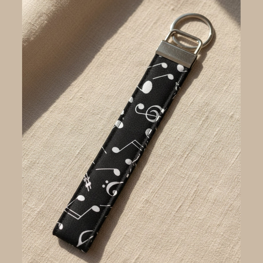A fabric wrist strap keychain featuring a black background with a white musical note pattern, finished with a silver metal split ring lying on a warm linen fabric