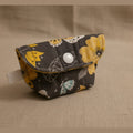 A small fabric pouch with a dark gray background and a yellow, white, and black floral pattern, featuring a silver snap button closure, resting on a light beige surface.