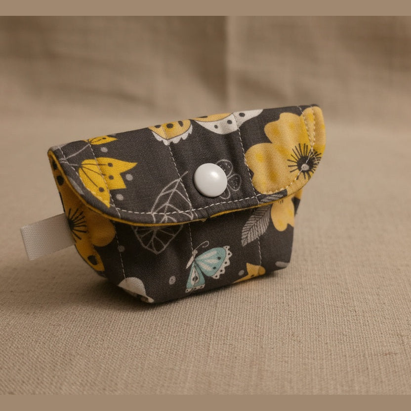 A small fabric pouch with a dark gray background and a yellow, white, and black floral pattern, featuring a silver snap button closure, resting on a light beige surface.