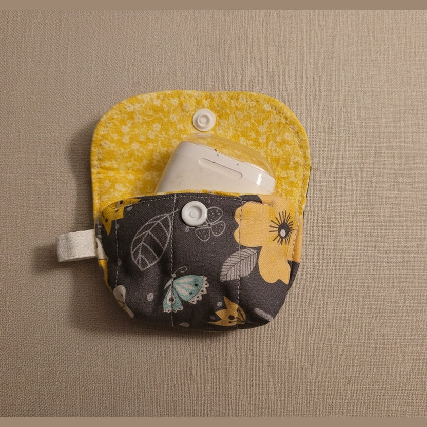 A small fabric pouch with a split design, featuring a yellow patterned top half and a dark gray floral pocket with yellow flowers, holding a white wireless earbud case, on a plain beige background.