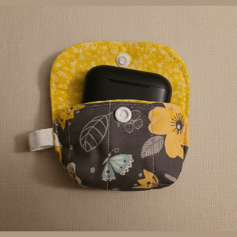 A small fabric pouch with a yellow floral top section and a dark gray lower section featuring butterfly and flower patterns, holding a black wireless earbud case. The pouch is photographed on a plain beige surface.