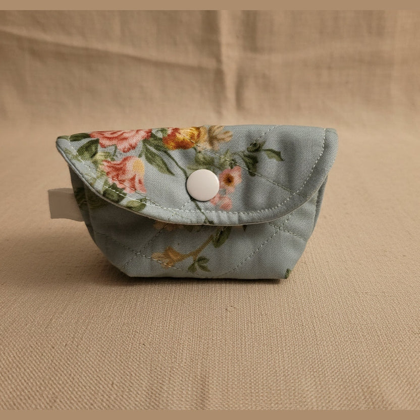 A small fabric pouch with a floral print in light blue, pink, and yellow, featuring a snap button closure, shown on a light beige surface.