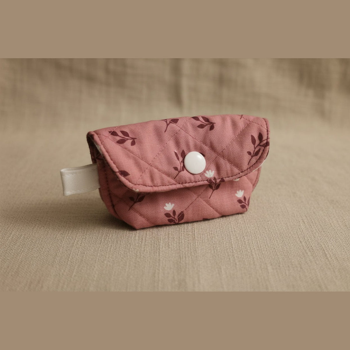 A small quilted fabric pouch in dusty rose pink with a dark floral pattern and a silver snap button closure, resting on a light-colored surface.