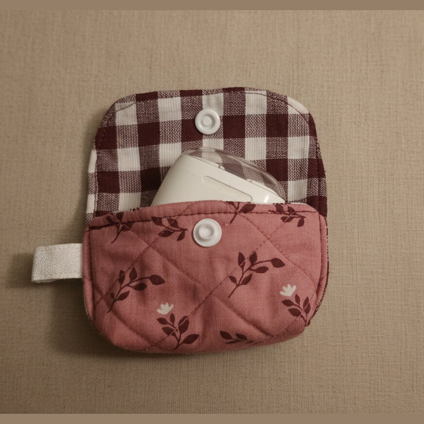 A small fabric pouch or case with a pink floral front pocket and a maroon-and-white gingham flap, shown on a plain beige background. A white item is partially tucked inside.