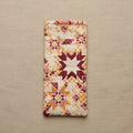 A rectangular quilted fabric pouch or cover with a geometric starburst pattern in cream, orange, and burgundy, hanging against a plain beige wall.