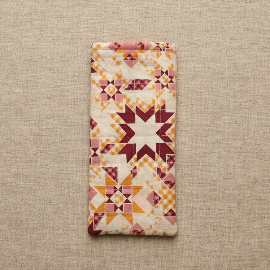 A rectangular quilted fabric pouch or cover with a geometric starburst pattern in cream, orange, and burgundy, hanging against a plain beige wall.