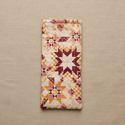 A rectangular quilted fabric pouch or cover with a geometric starburst pattern in cream, orange, and burgundy, hanging against a plain beige wall.