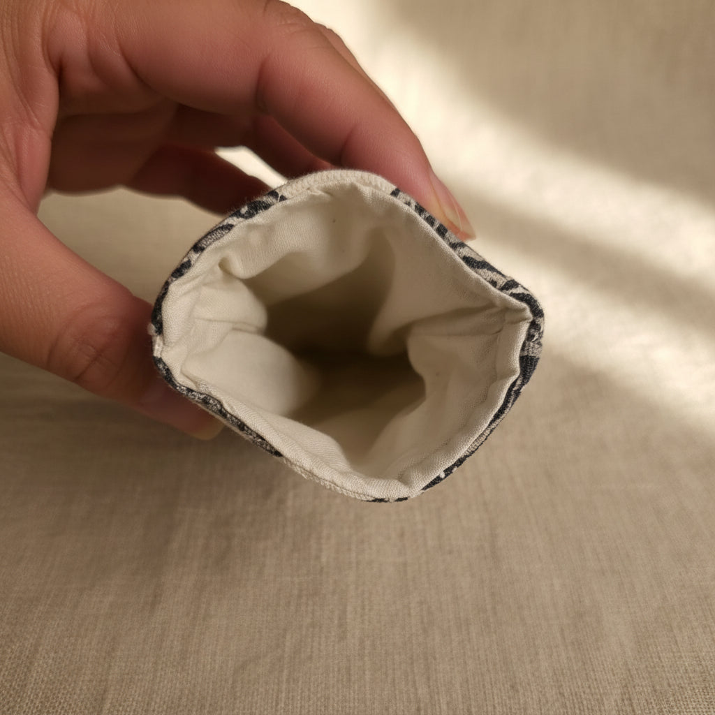 A close-up of a hand holding the open end of a small fabric pouch or sleeve, showing a cream-colored fabric lining and a dark patterned outer fabric.