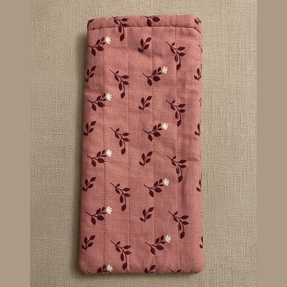 A pink quilted fabric pouch or sleeve with a dark floral pattern, hanging on a plain beige wall above a gray textured surface.