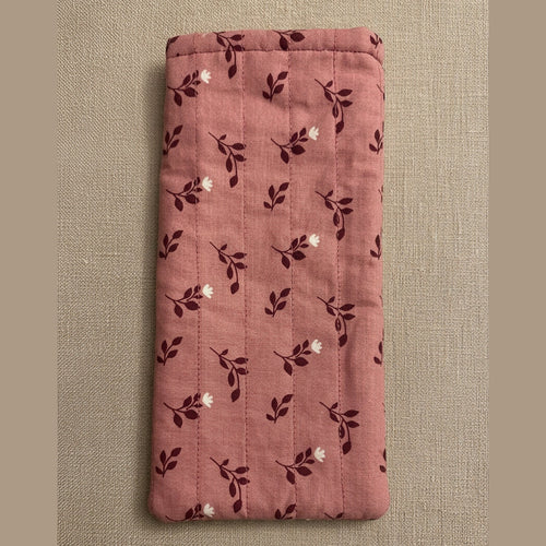 A pink quilted fabric pouch or sleeve with a dark floral pattern, hanging on a plain beige wall above a gray textured surface.