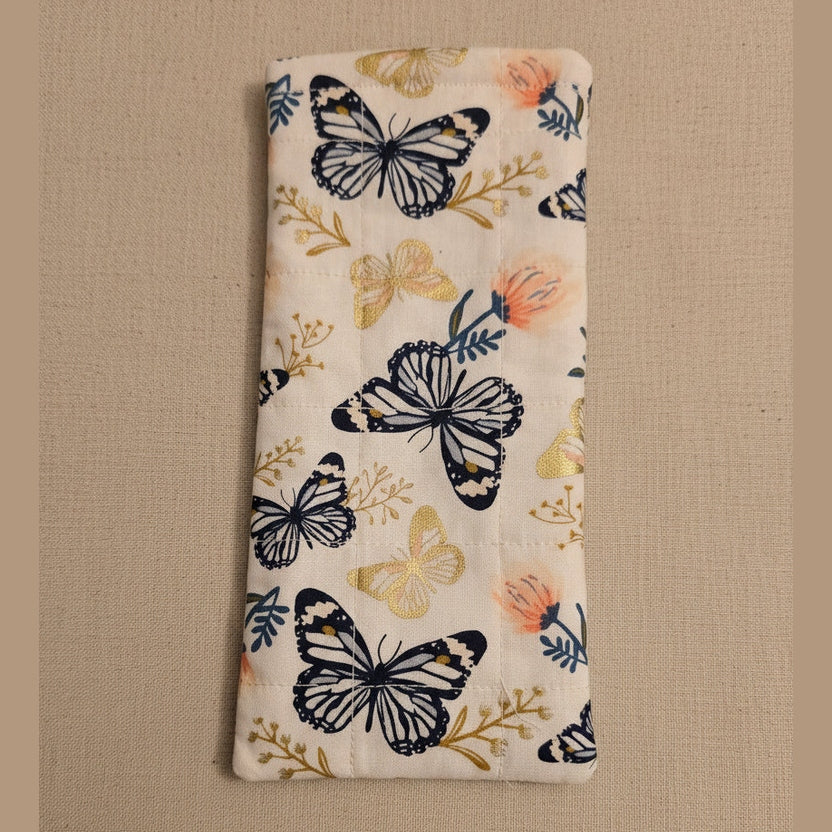 A rectangular quilted fabric pouch with a printed pattern of black and gold butterflies and small peach flowers on a cream background, shown against a plain beige wall.