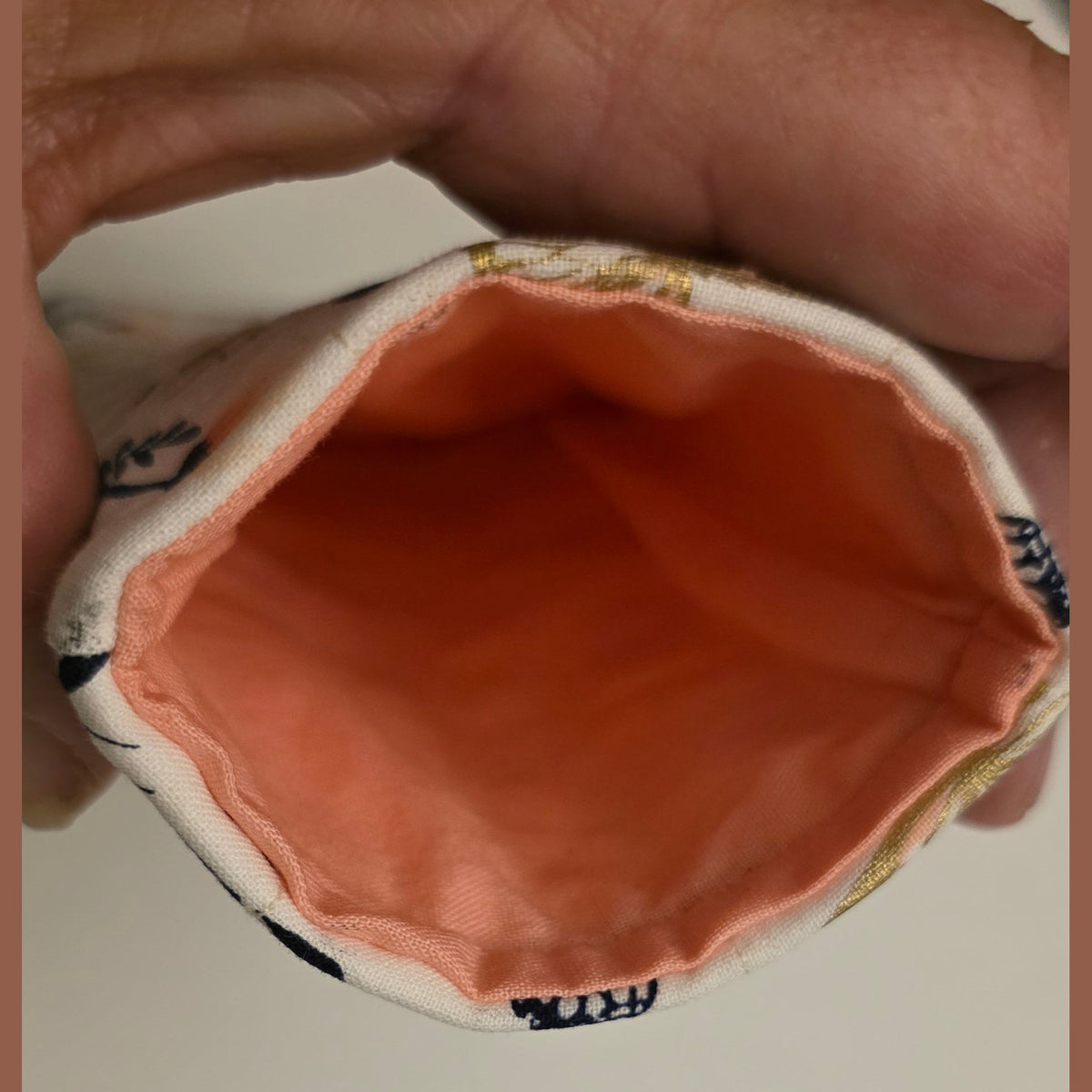 A close-up of a hand holding the open end of a small fabric pouch or bag, showing a peach-colored interior lining and a patterned exterior edge.