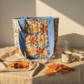 A fabric tote bag with a colorful floral print and solid blue handles, standing on a beige fabric-covered surface