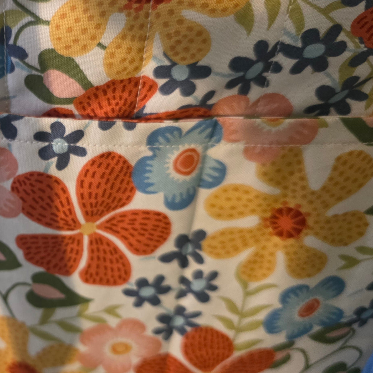 Close-up of a fabric tote bag showing a blue denim-like side panel and a floral front pocket with orange, blue, yellow, and white flowers on a cream background.