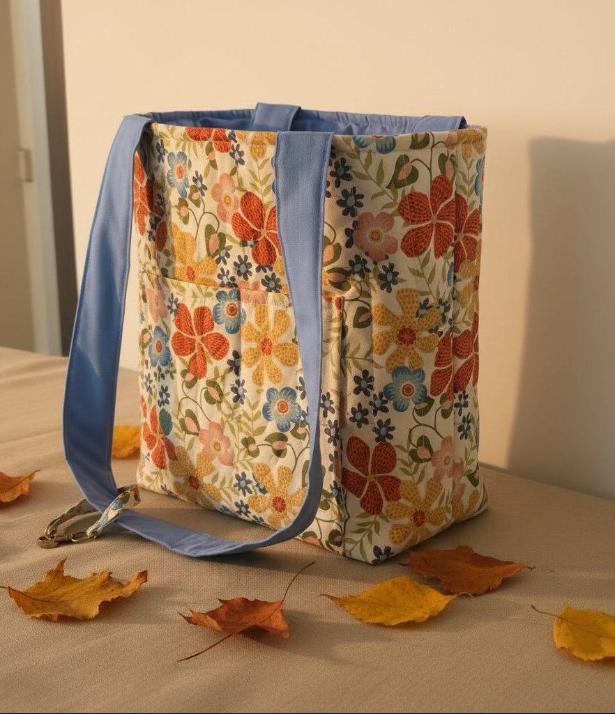 A tall fabric tote bag side view with a colorful floral print and blue handles, resting on a light-colored cushion
