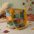 A handmade quilted tote bag with a multicolor patchwork pattern of orange, blue, green, and yellow fabrics, featuring a solid mustard yellow handle and trim, sitting on a beige fabric-covered surface.