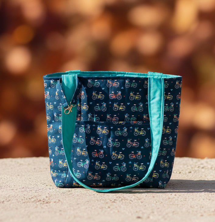 A fabric tote bag with a dark navy blue background covered in a colorful bicycle pattern, featuring bright teal-green trim and handles, sitting on a beige fabric surface.