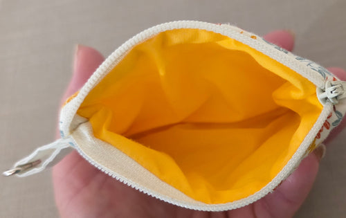 A close-up of a small fabric zip pouch held in a hand, showing a bright yellow interior lining and a cream zipper with a small floral print on the exterior.