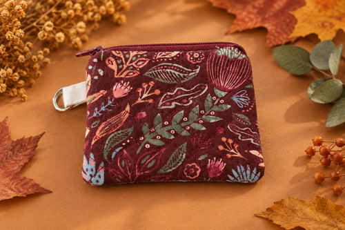 A small fabric zippered pouch with a floral and botanical pattern in burgundy, pink, green, and blue, featuring a white plastic clip at the top, shown on a textured beige fabric background.