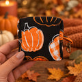A hand holding a small fabric pouch with a black background and orange pumpkin pattern, featuring an orange zipper pull with a metal clasp.