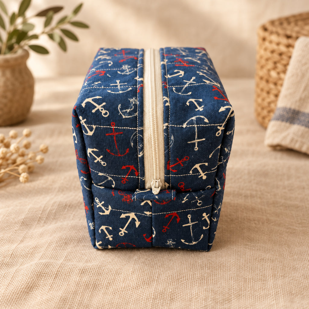 A square zippered fabric pouch with a navy blue background and a white, red, and blue anchor pattern, resting on a beige fabric surface.