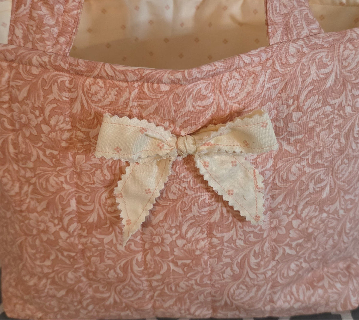 A small fabric tote bag with a dusty pink and white damask pattern, featuring a cream-colored fabric bow on the front and a cream lining inside.