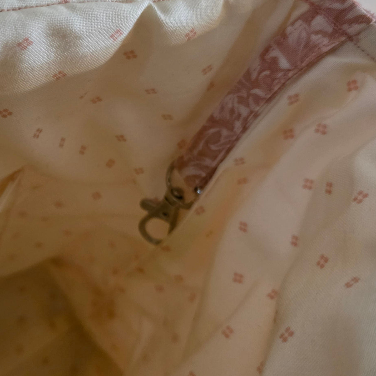 Close-up of the interior lining of a soft bag or pouch, showing a cream fabric with a small pink floral pattern and a metal lobster clasp.