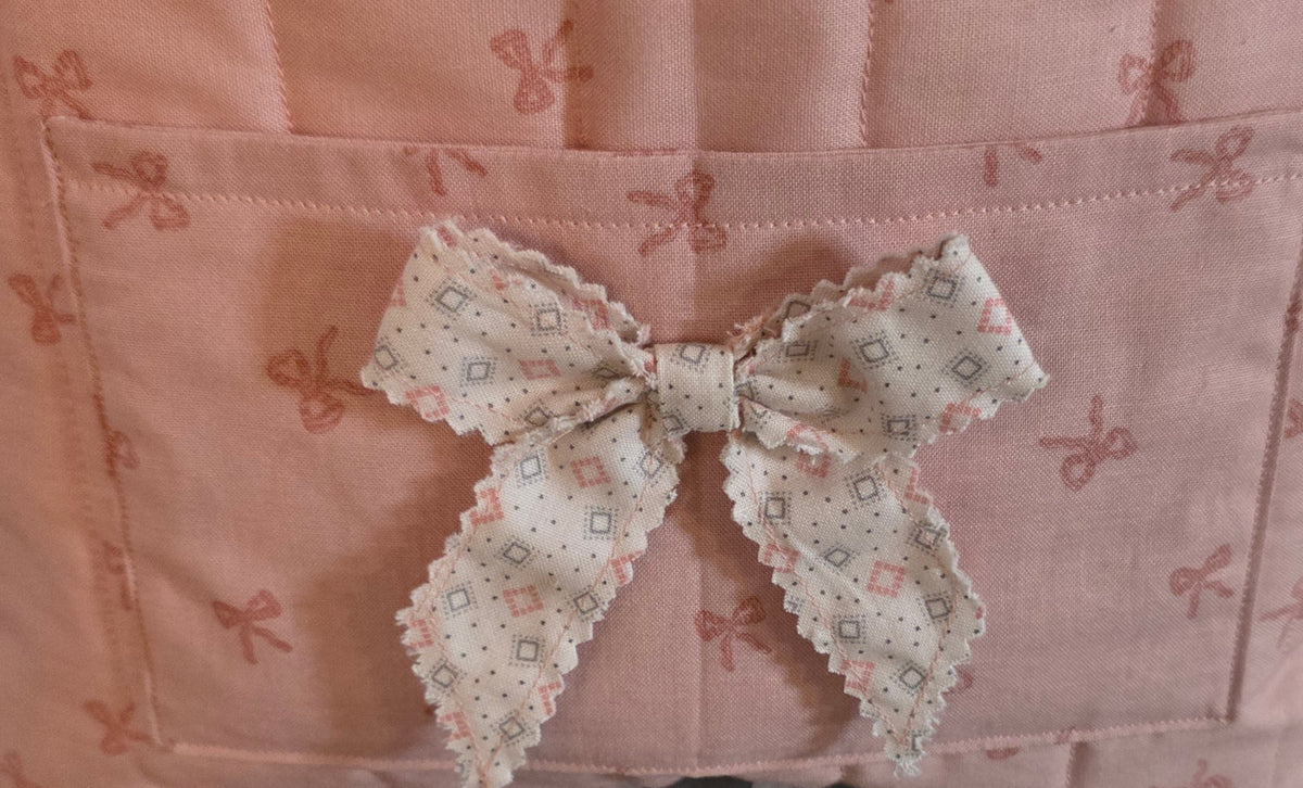 A close-up of a pink quilted fabric organizer or basket with a front pocket and a decorative bow made from a small floral print fabric.
