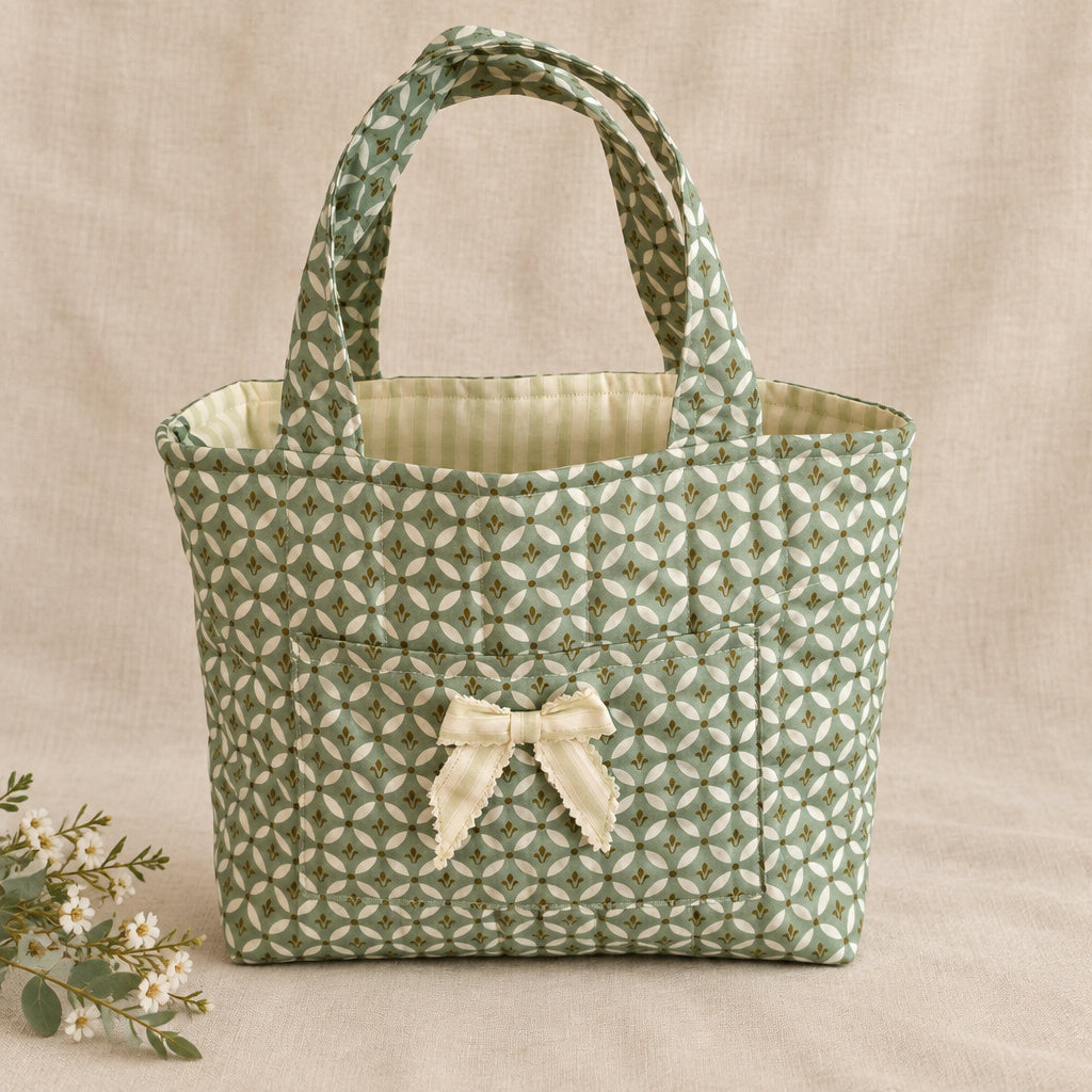 A small fabric tote bag with a green and white geometric pattern, dual top handles, and a cream-colored bow on the front.