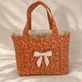 An orange fabric tote bag with a small white floral pattern, featuring dual top handles and a decorative white bow on the front pocket